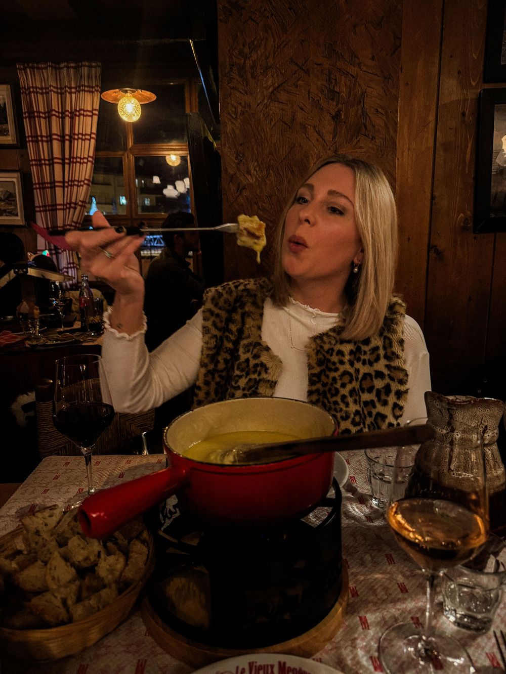 Blonde woman eating cheese fondue at a cozy alpine restaurant wearing a leopard print vest.
