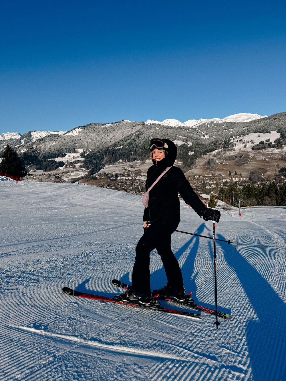 Woman skiing on groomed alpine slope with scenic mountain village and blue sky in background.