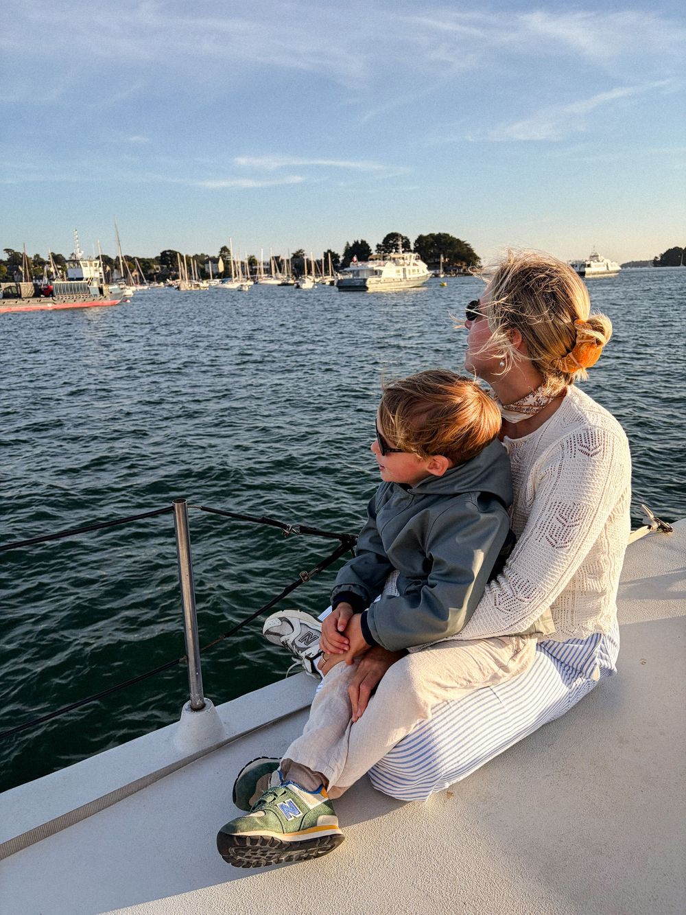 Adult and toddler sitting on boat bow, overlooking calm marina with sailboats at golden hour.