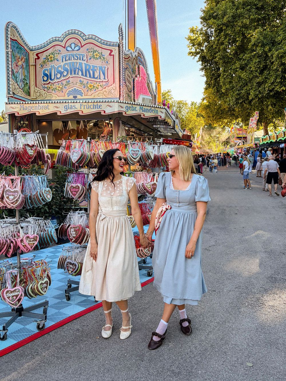 Two women in vintage dresses pose at Oktoberfest near a Süsswaren candy stall with heart-shaped gingerbread decorations.