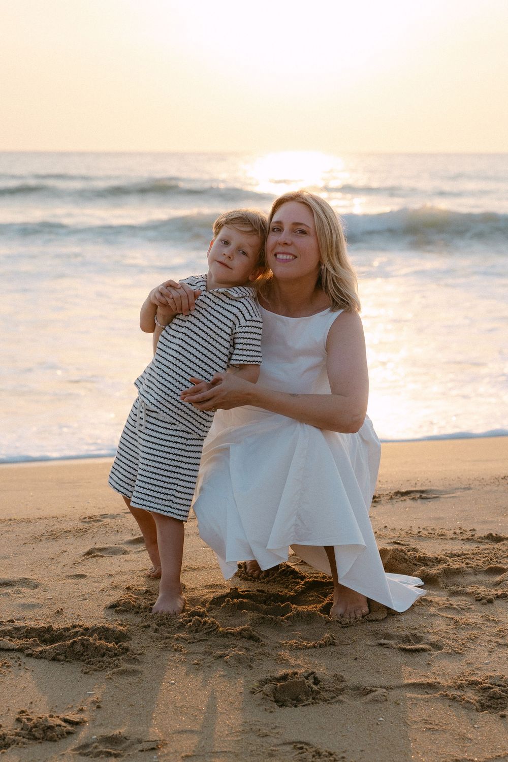 A mother in a white dress hugs her young child on a sandy beach at sunset.