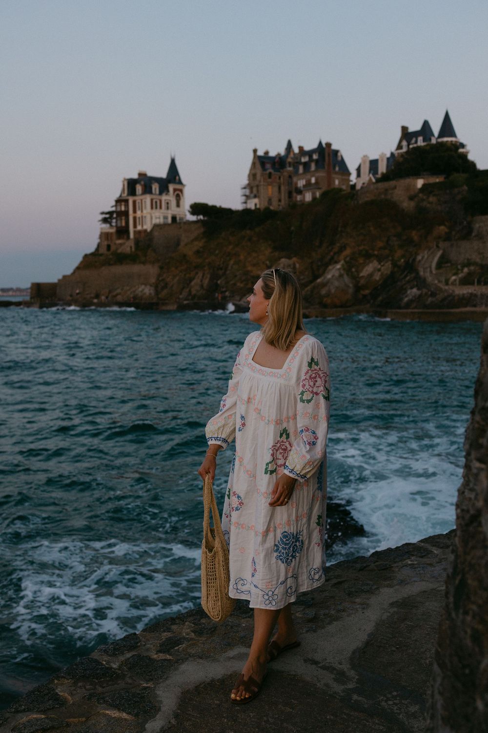 Woman in floral dress holding woven bag stands on rocky coast with historic cliffside mansion at dusk.