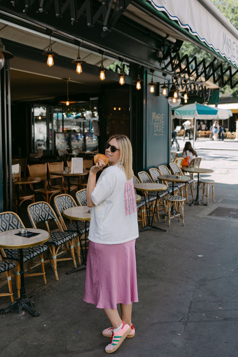 Woman in white tee and pink skirt eating a croissant outside a Parisian cafe with bistro chairs.