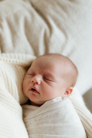 A sleeping newborn baby wrapped in soft white knit blankets lays peacefully against white bedding.