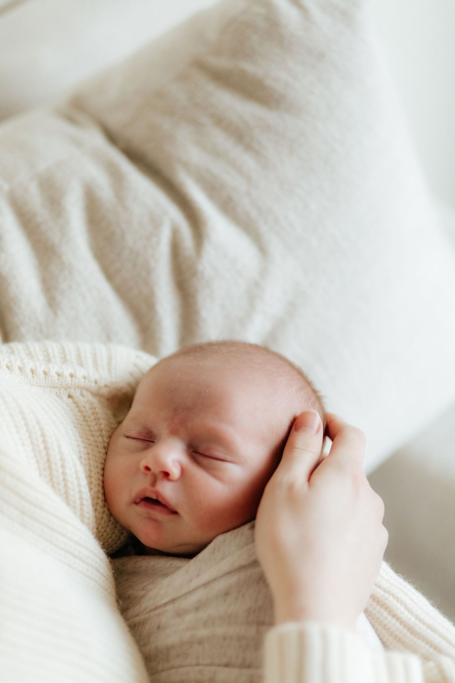 A newborn baby peacefully sleeping on white knitted fabric while a gentle hand caresses their cheek.