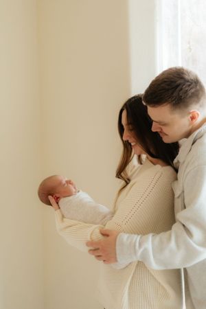 A tender family moment as parents in cozy sweaters hold their newborn baby against a light backdrop.