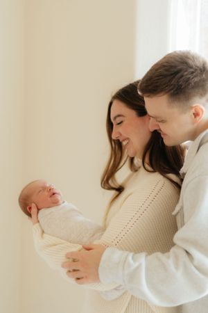 Parents in white sweaters share a joyful moment with their newborn baby against a light background.
