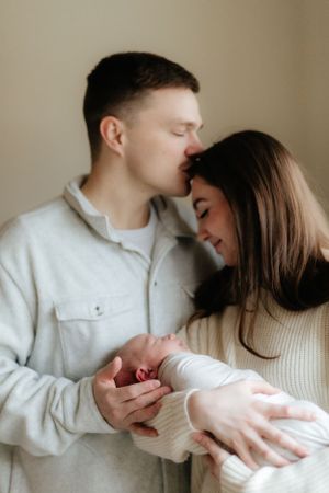 Intimate portrait of a couple in white sweaters sharing a loving moment while cradling their newborn baby.