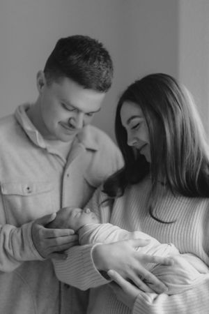 A tender black and white portrait of a couple lovingly gazing at their newborn baby wrapped in white.