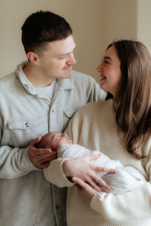 New parents share an intimate moment with their newborn baby dressed in white while standing against a neutral wall.