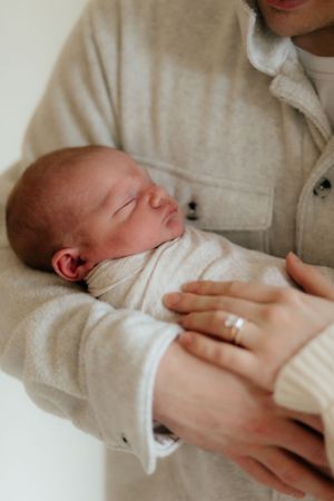 A sleepy newborn baby dressed in white rests peacefully in caring arms with a gold ring visible.
