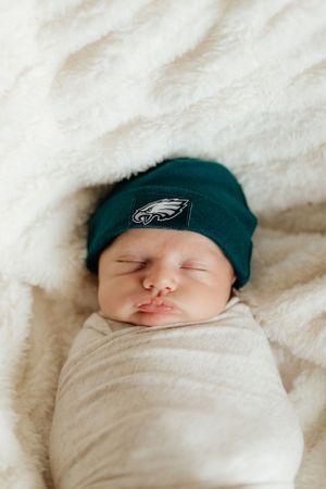 Sleeping newborn wrapped in white swaddle blanket wearing Philadelphia Eagles green knit beanie hat.