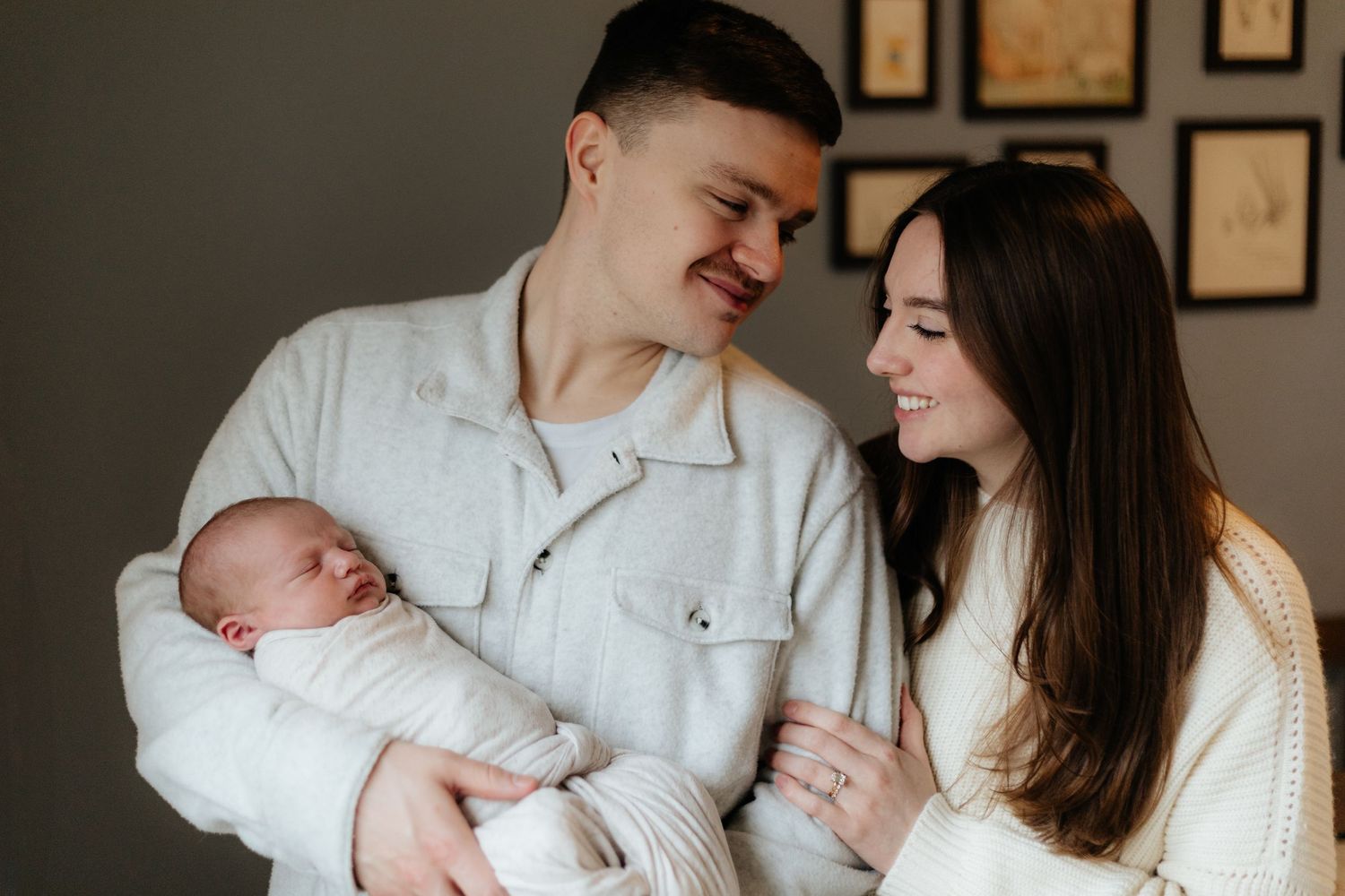 A couple looks lovingly at their newborn baby while standing in front of framed wall art.