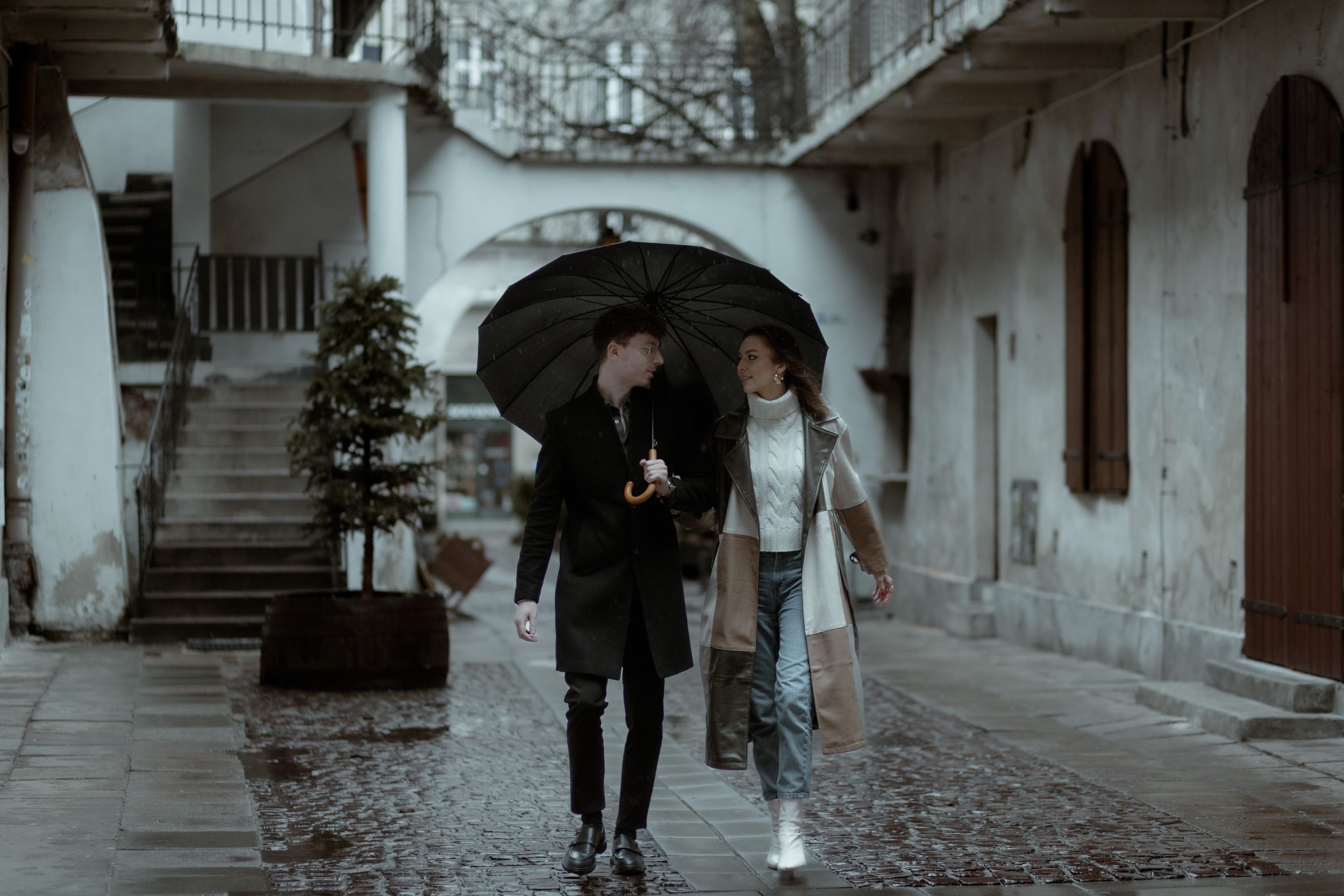 A couple walking together with a black umbrella in a cobblestone courtyard with arches and stairs.
