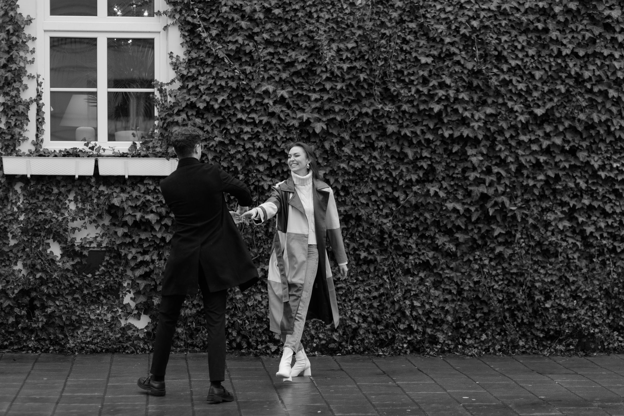 Two people in black and white photography standing against an ivy-covered wall near a window.