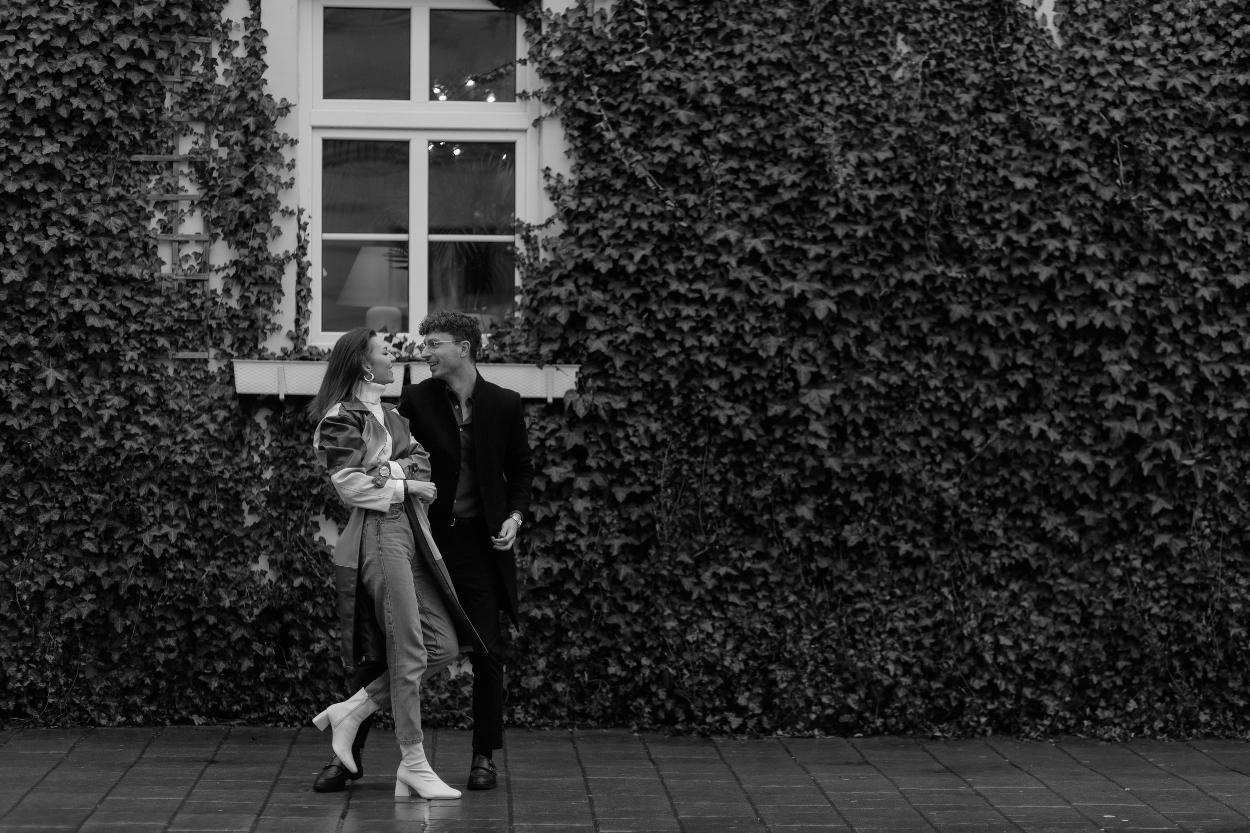 A couple shares a romantic moment against an ivy-covered wall with a window in black and white photography.