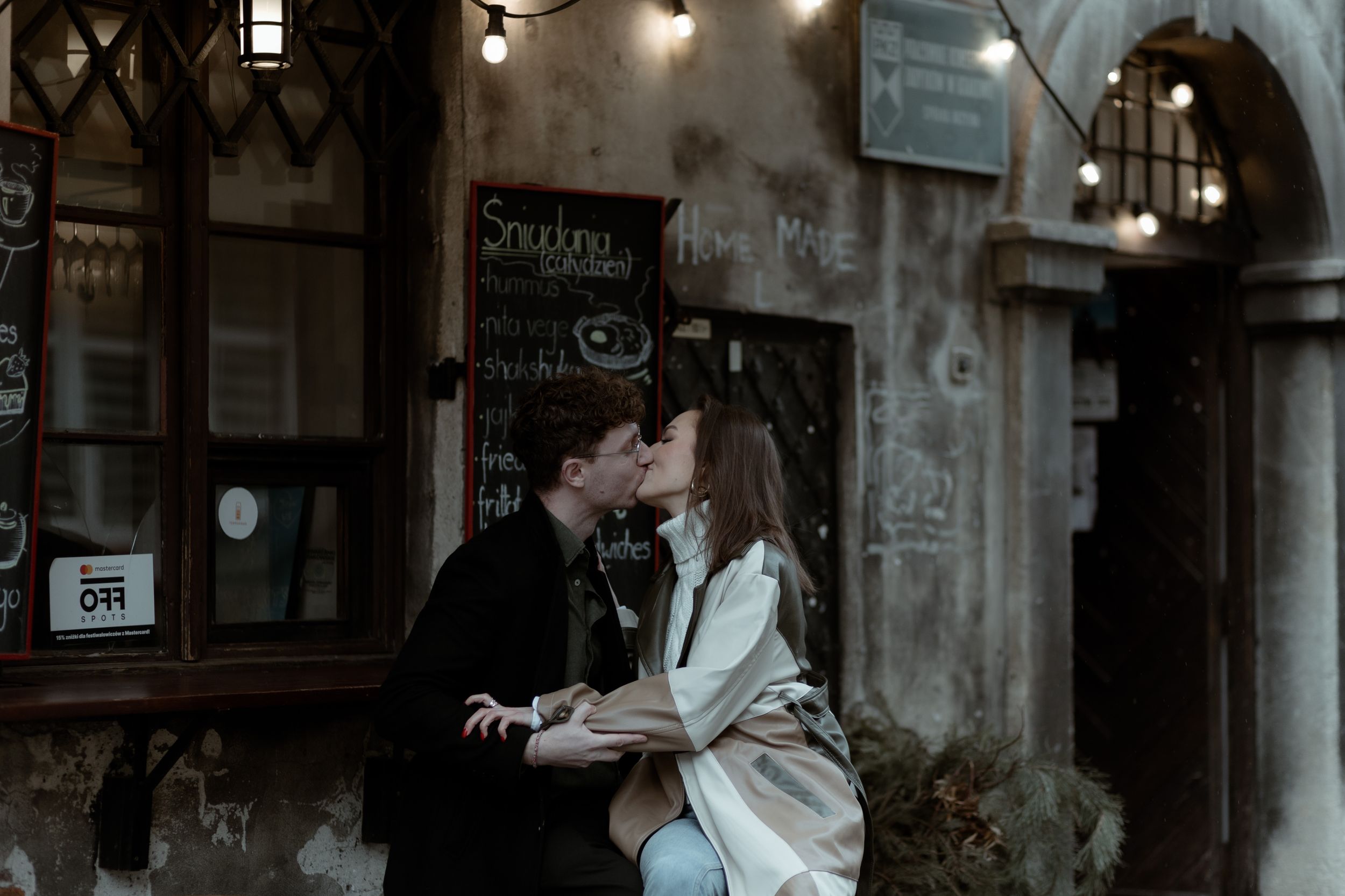 A romantic couple embraces in front of a vintage storefront with moody lighting and classic architecture.