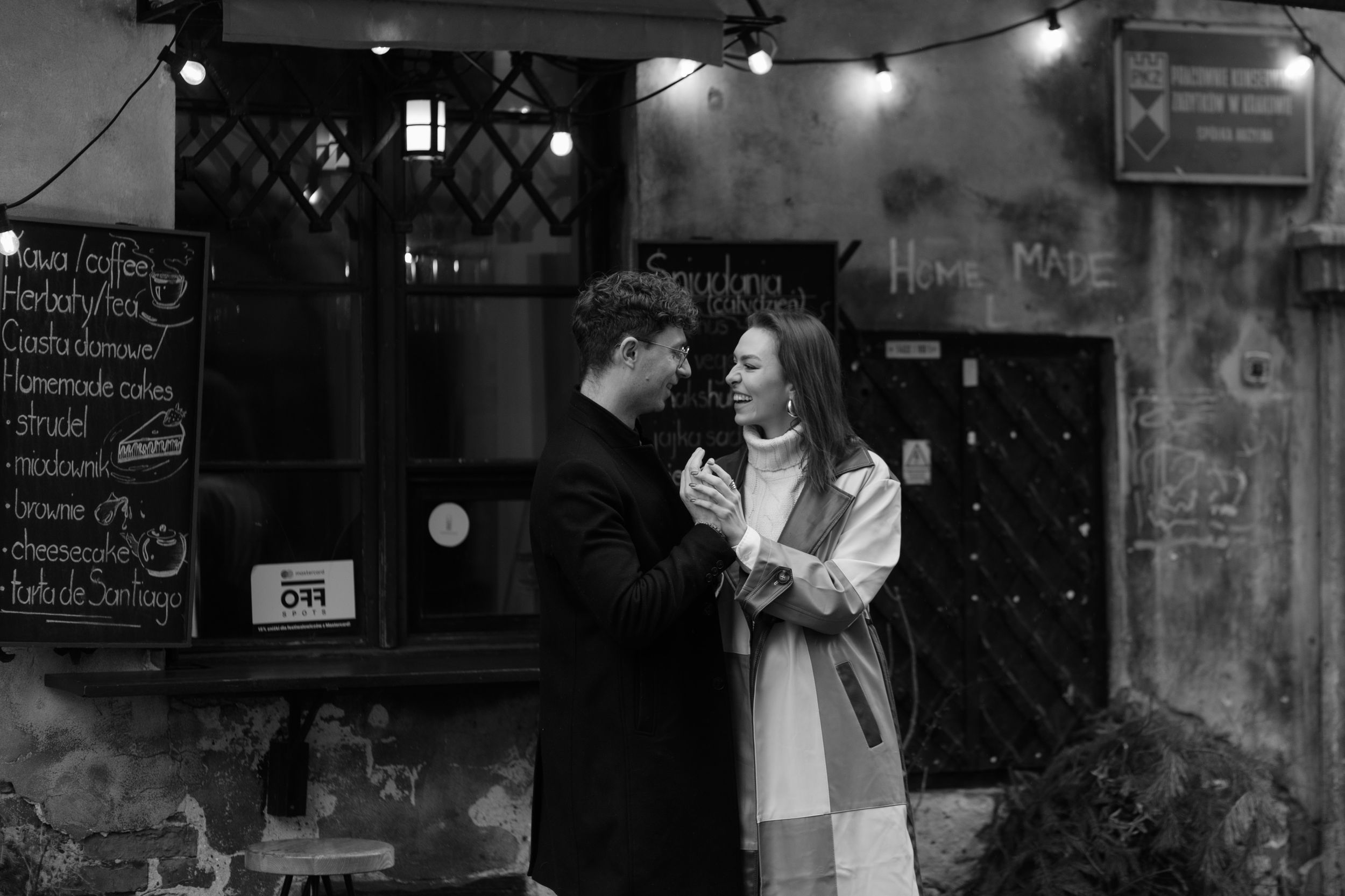Black and white photo of a couple in casual winter outfits sharing a romantic moment under string lights outside a cafe.