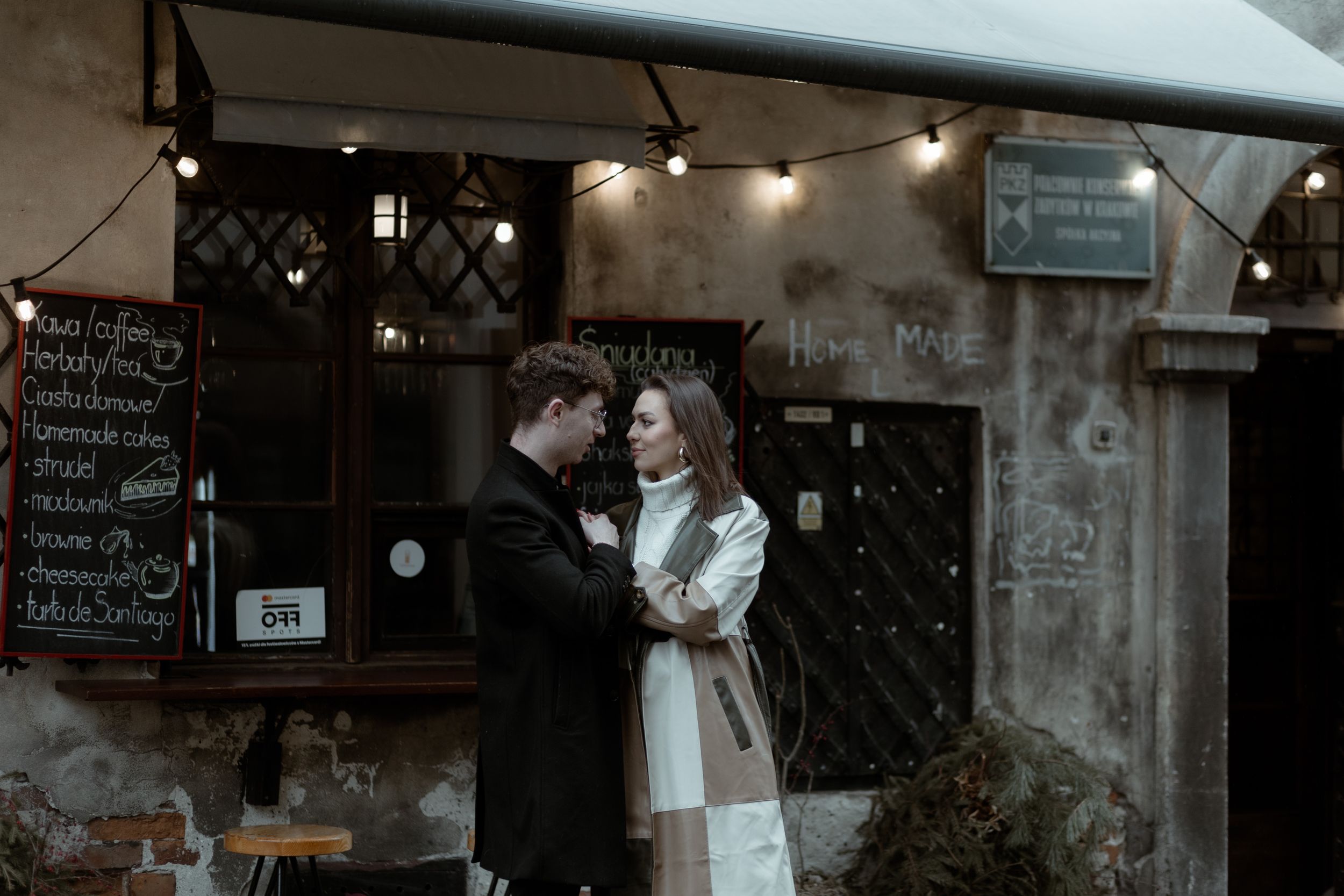 A romantic couple embraces in front of a rustic cafe with string lights and chalkboard menu at dusk.