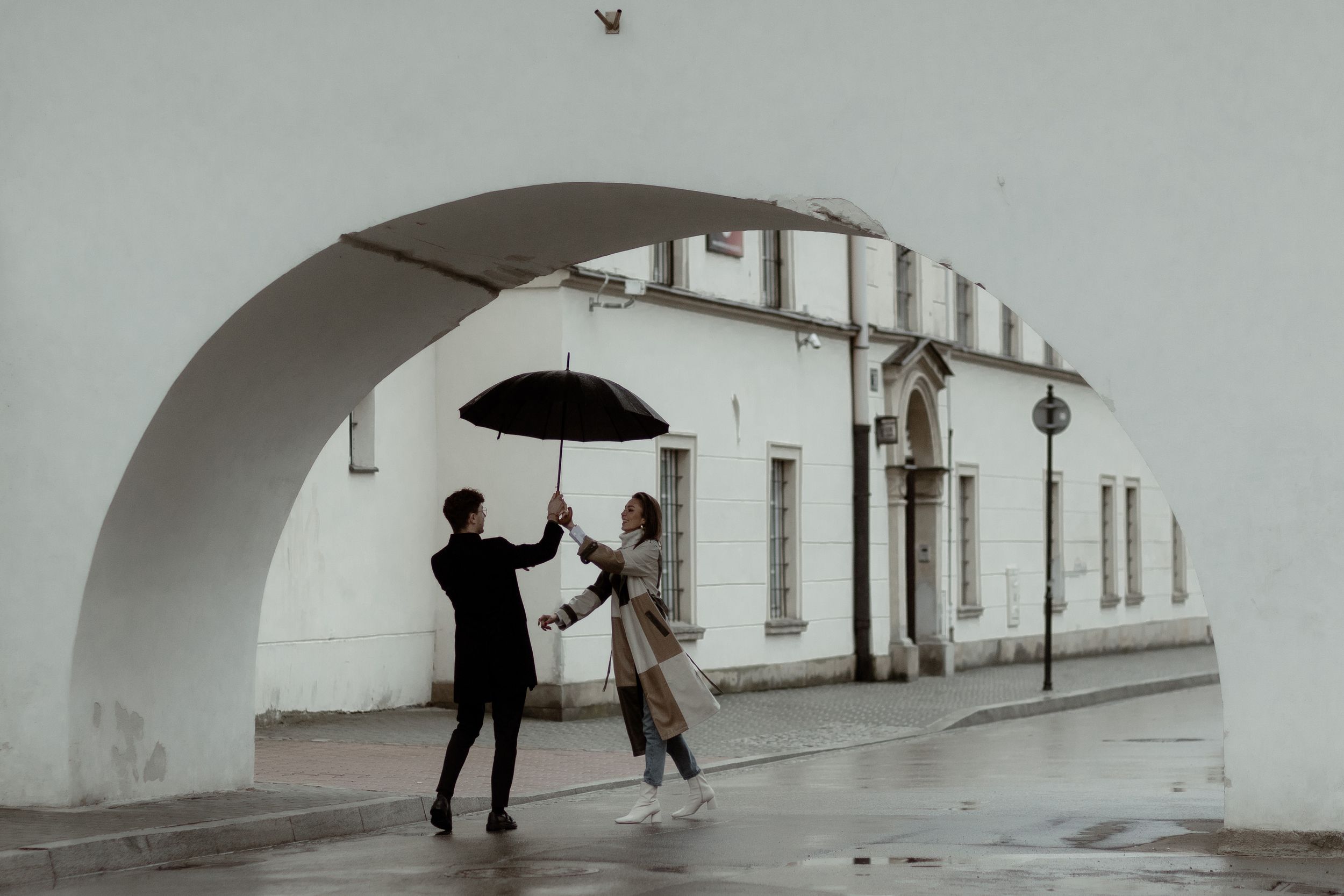 Silhouettes of two people dancing joyfully under an archway as one holds an umbrella on a rainy street.
