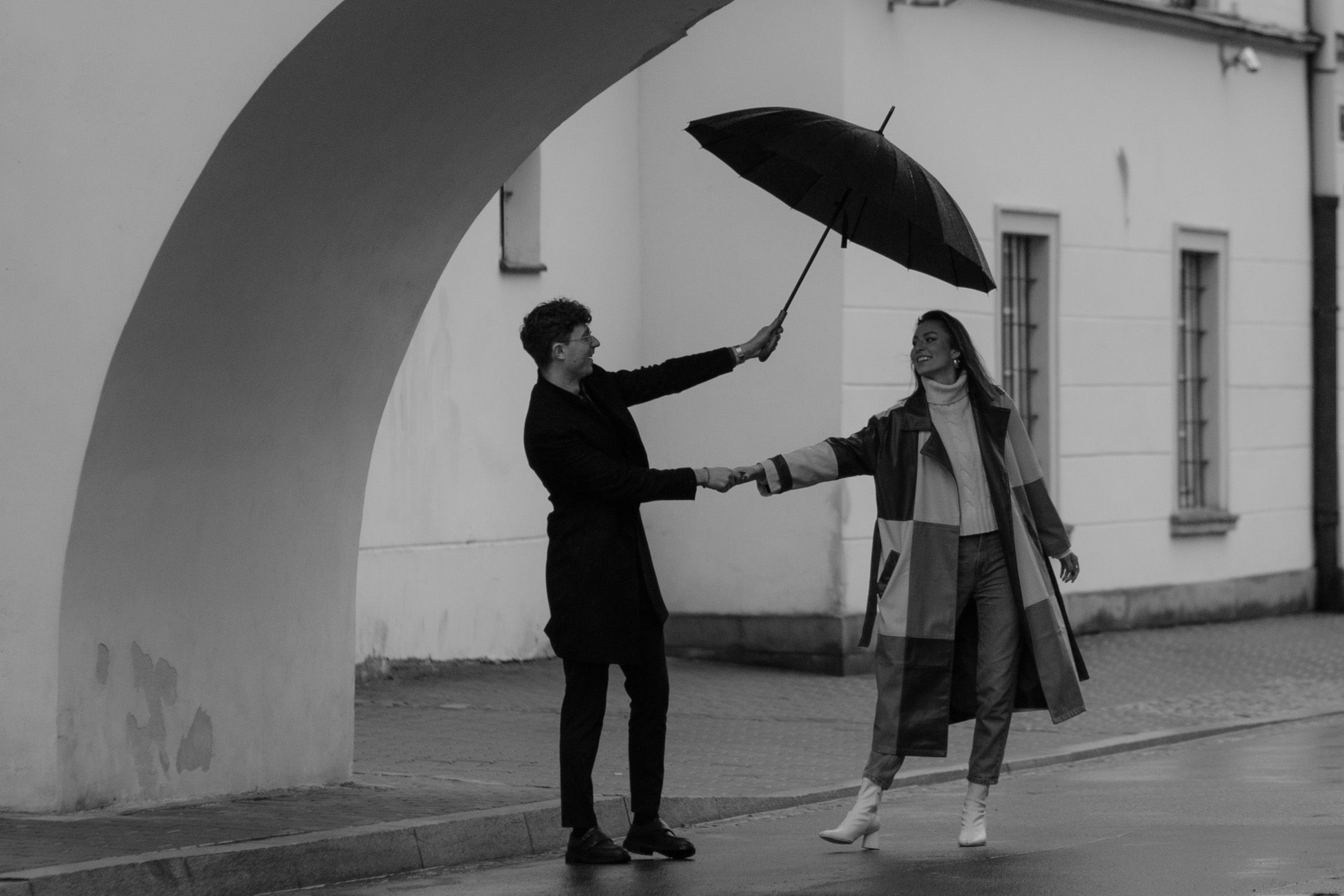 A romantic black and white scene showing two people under an archway with one holding an umbrella.