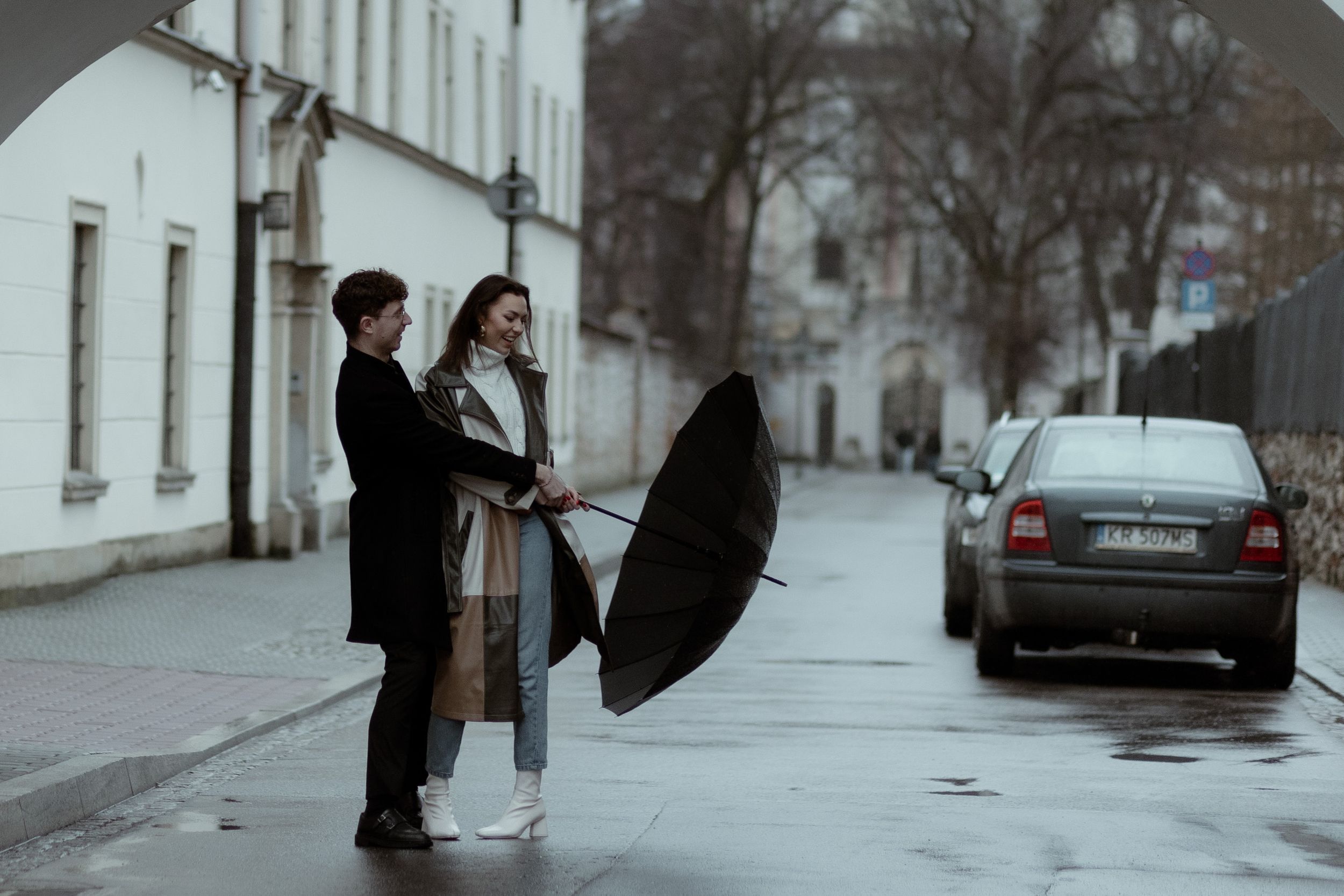 Two people stand together on a rainy city street with a black umbrella and parked car in the background.