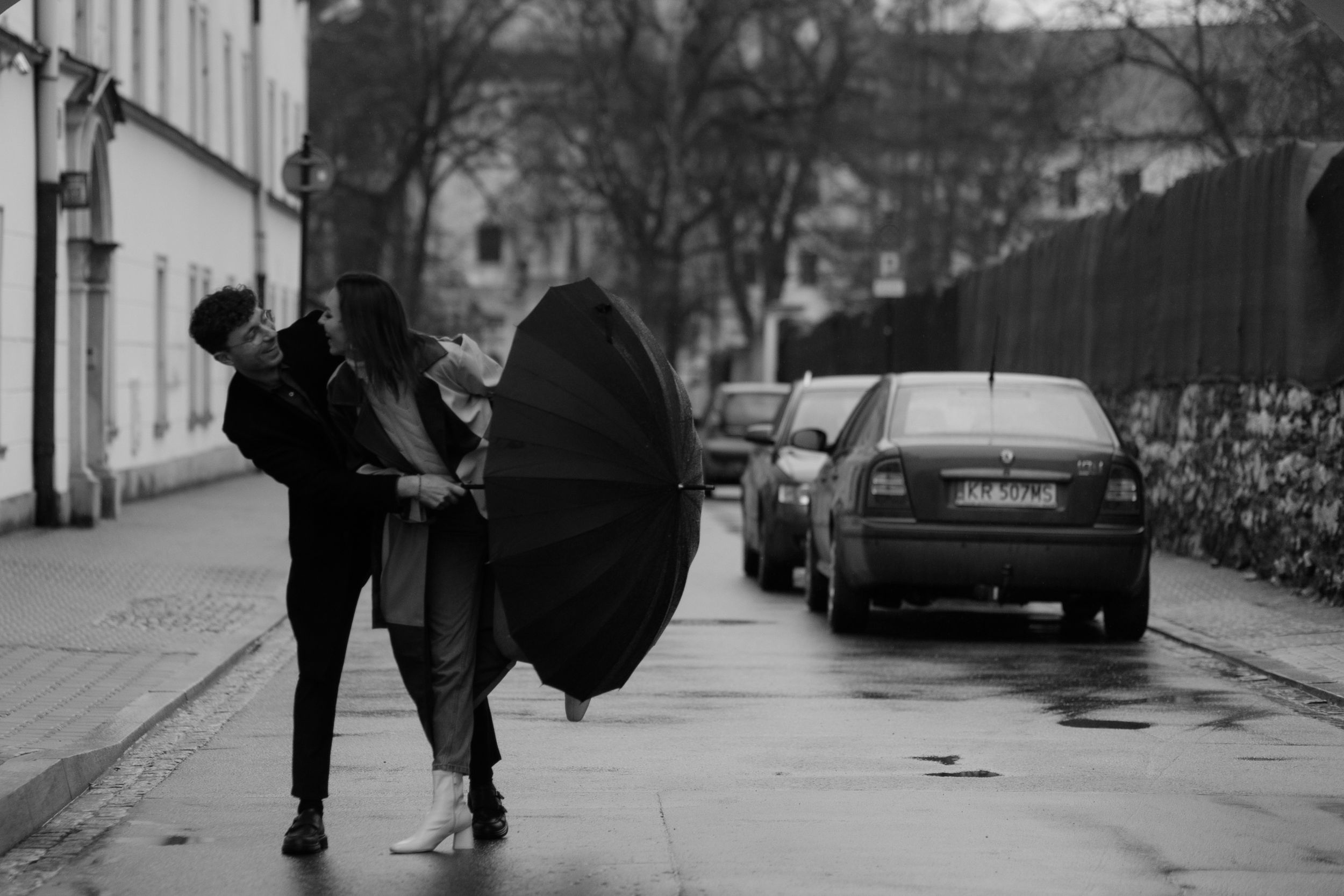 A figure struggles with an umbrella being blown by strong wind on a black and white urban street.