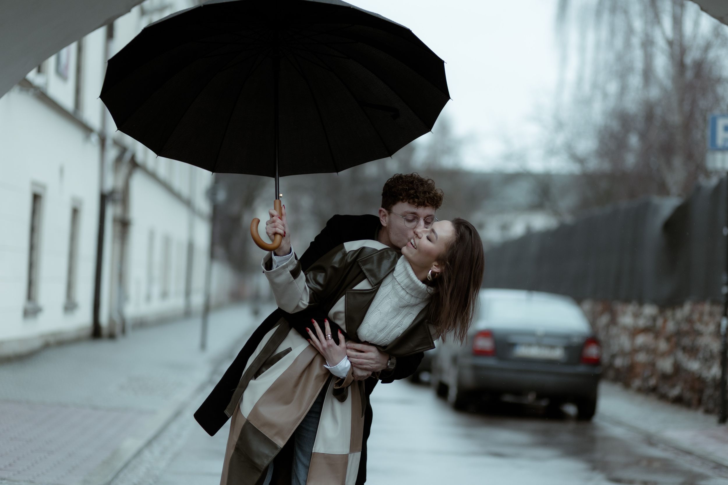 Person in beige coat struggles with inverted black umbrella on a windy urban street.