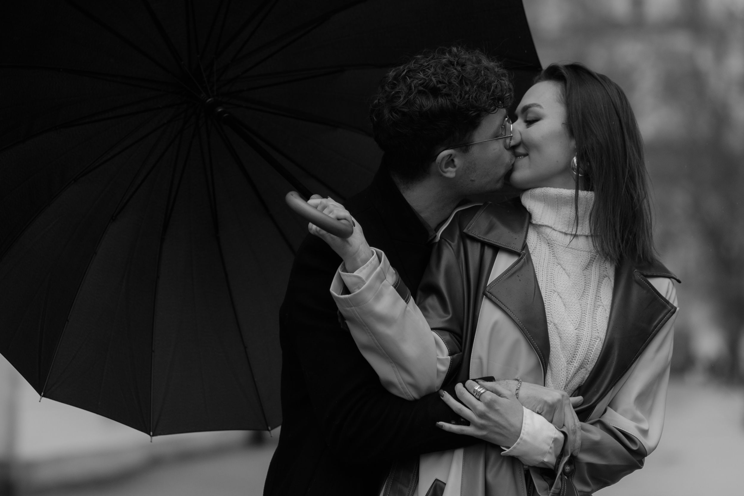 A couple shares an intimate moment under a black umbrella in a romantic black and white photograph.