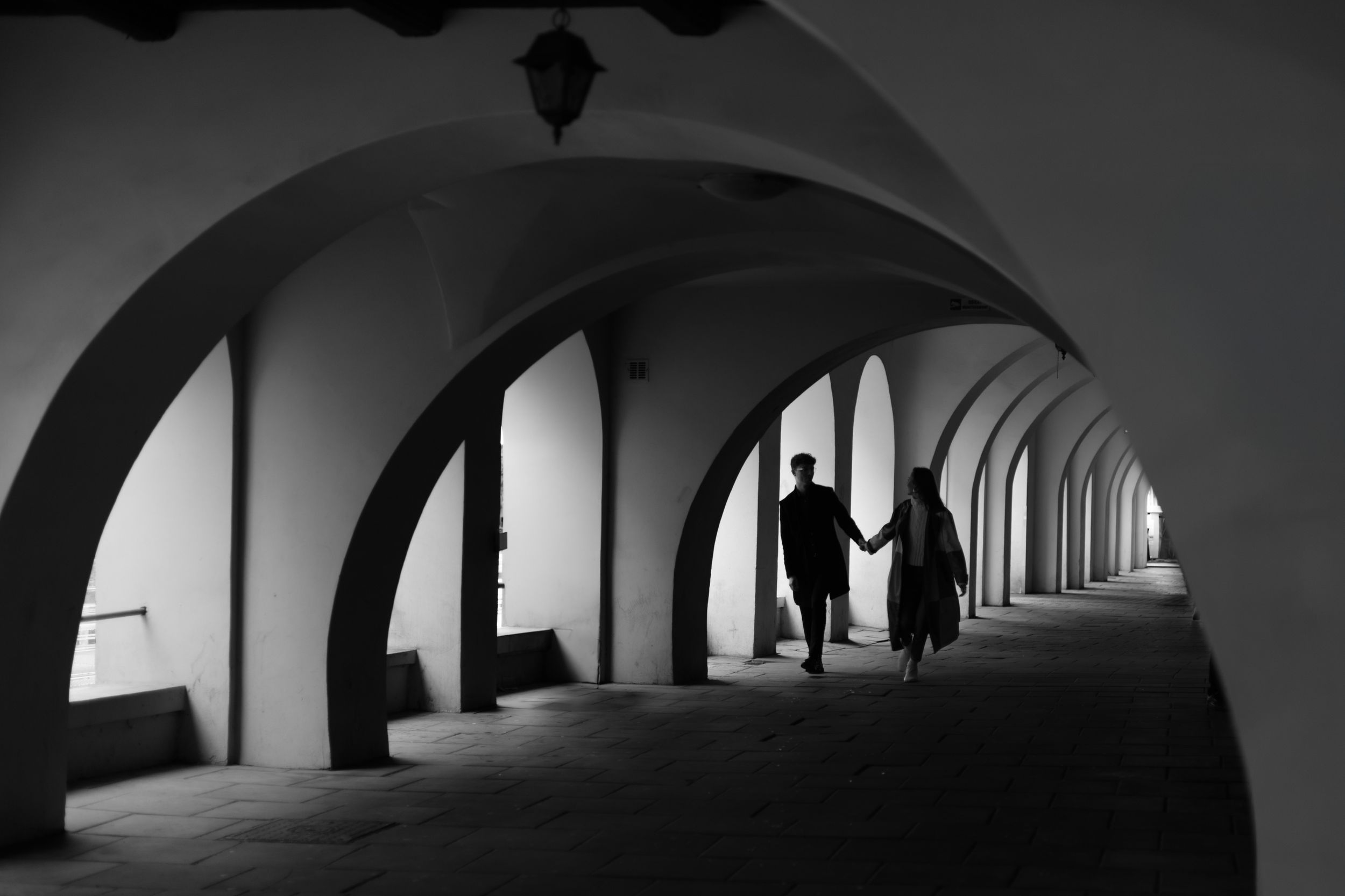 Black and white architectural photograph showing silhouetted figures in a repeating arched colonnade hallway.