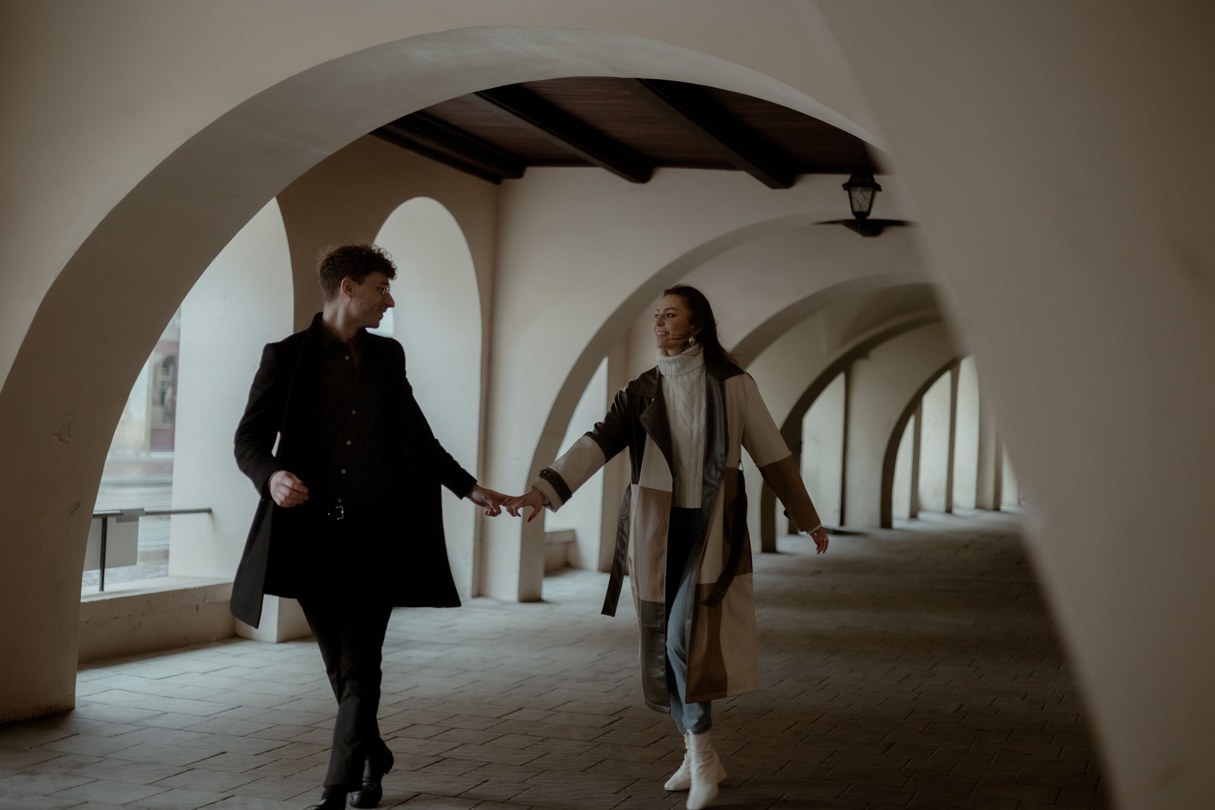 Two people holding hands while walking through an arched corridor with a playful and joyful mood.