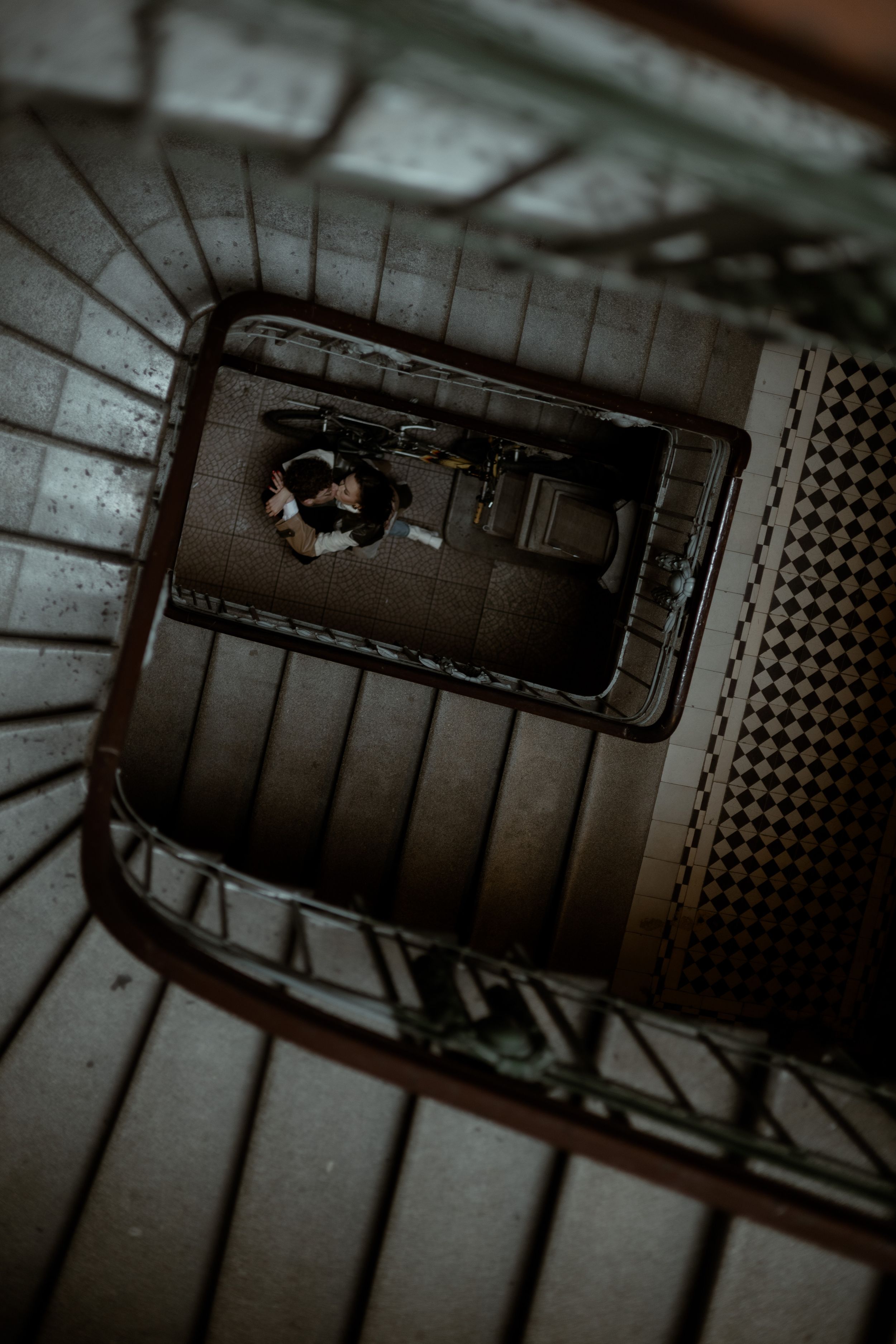 Looking down a dark spiral staircase in an old building with dim lighting and metal railings creating dramatic shadows.