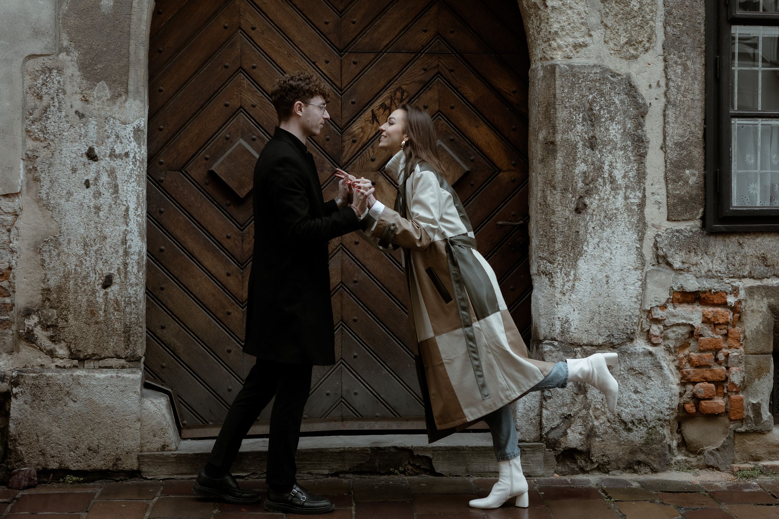 Two people in vintage style clothing dance together in front of a rustic wooden door against a weathered stone wall.
