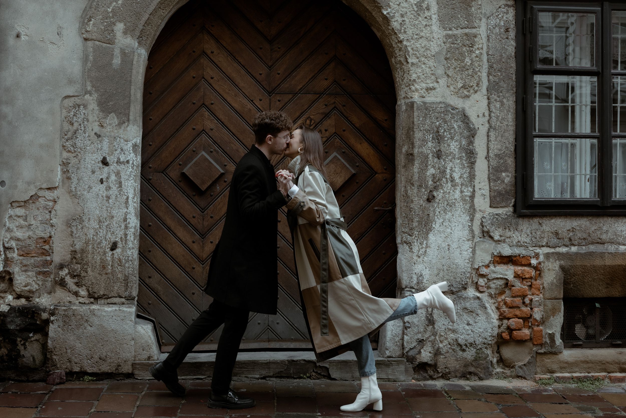 A romantic vintage-styled couple embraces in front of a weathered arched wooden door and brick wall.