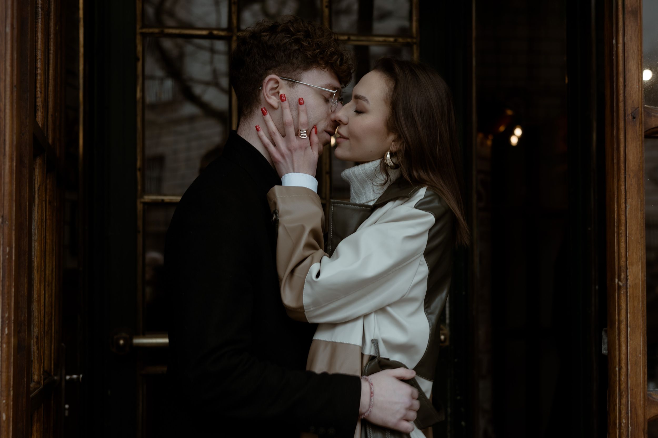 Couple shares an intimate kiss in a shadowy doorway while dressed in elegant winter attire.