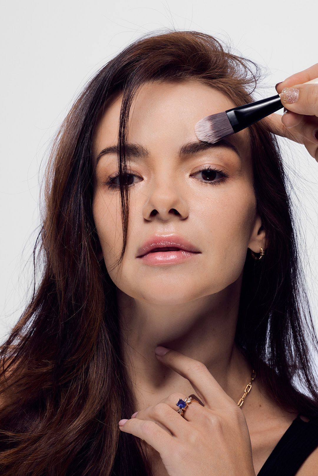 In a beauty portrait, a makeup brush applies eyeshadow while the subject wears a black top and gemstone ring.