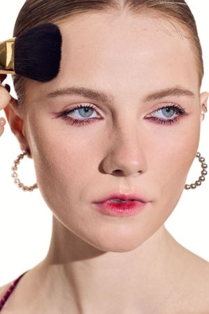 Makeup application with brush against light background showing pink eyeshadow and coral lipstick.