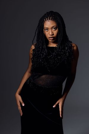 Studio portrait against dark background of a person wearing a black dress with sheer details and long braided hairstyle.