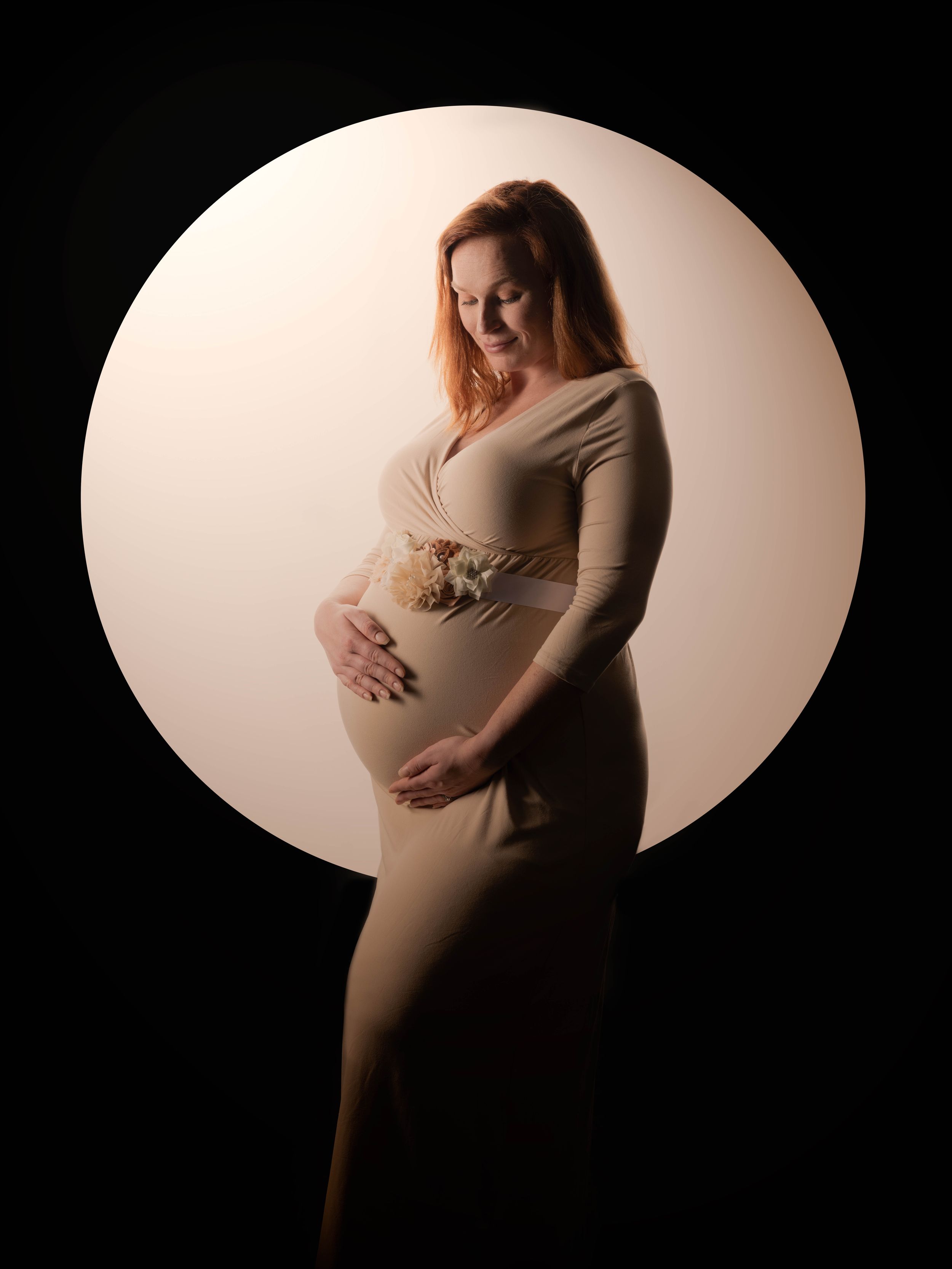 A pregnant woman in a beige dress poses against a circular backdrop with dramatic lighting for a maternity photoshoot.