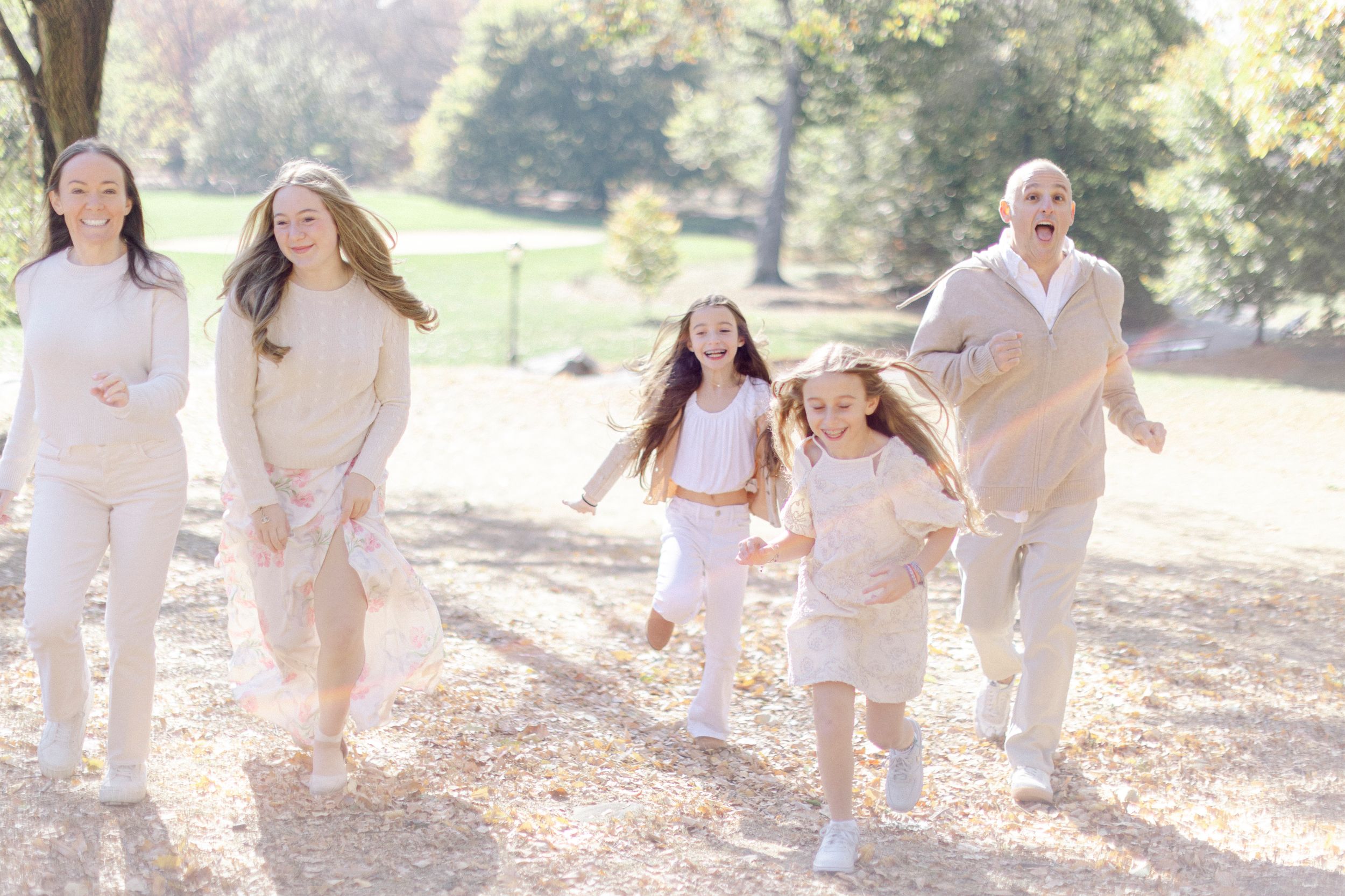 Family in light neutral colored outfits running and playing together in a sunny outdoor park setting.