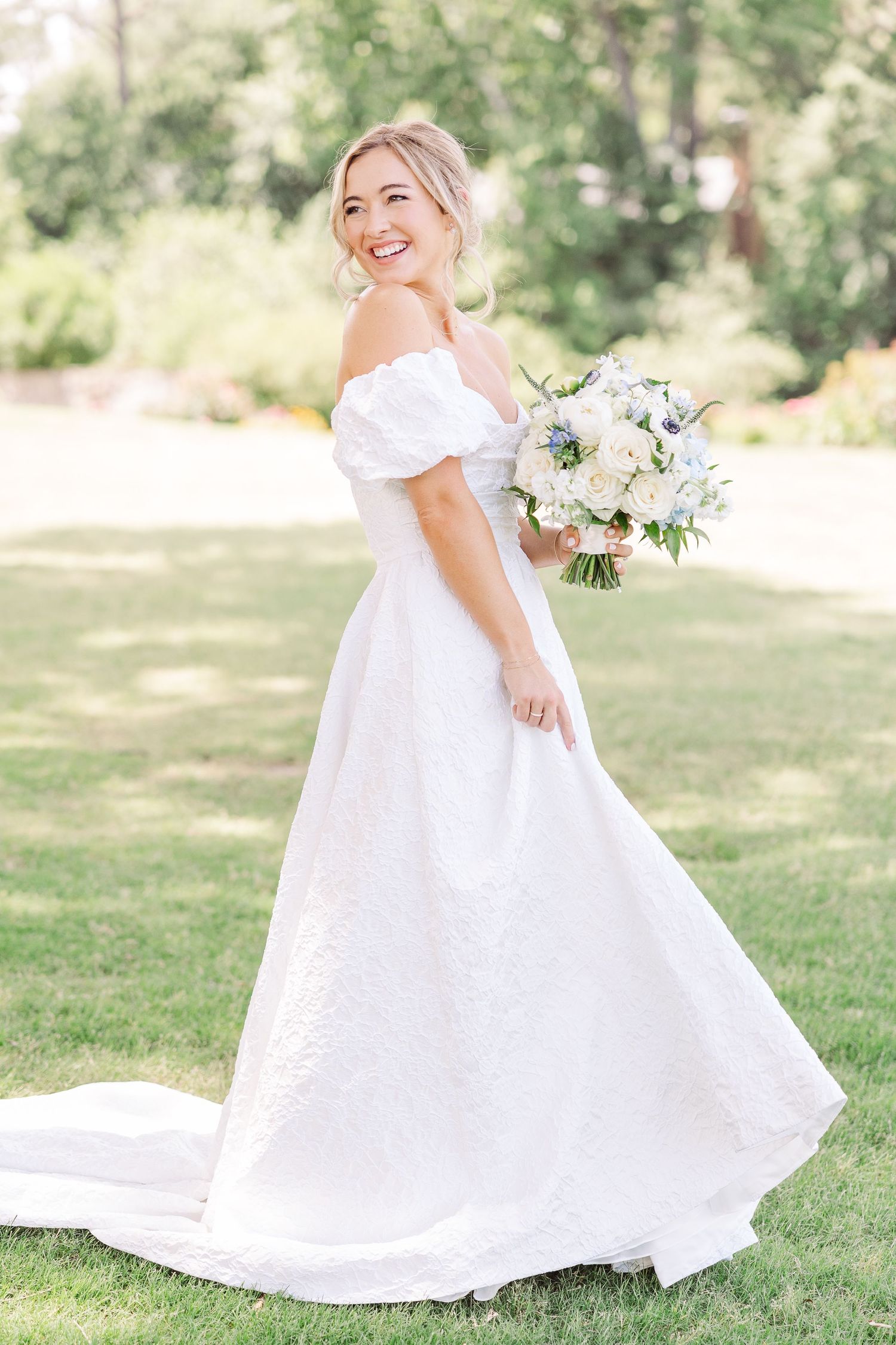 A bride in an elegant off-shoulder white wedding gown smiles while holding a pastel bouquet in a sunny outdoor garden setting.