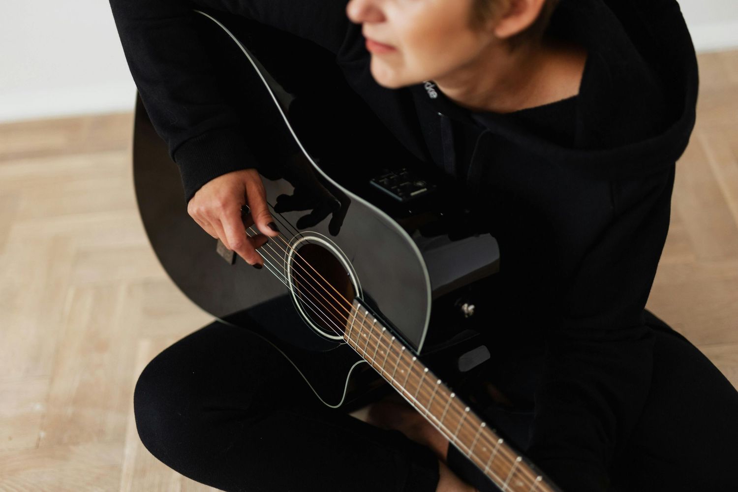A musician in a black outfit sits with an acoustic guitar on a wooden floor.