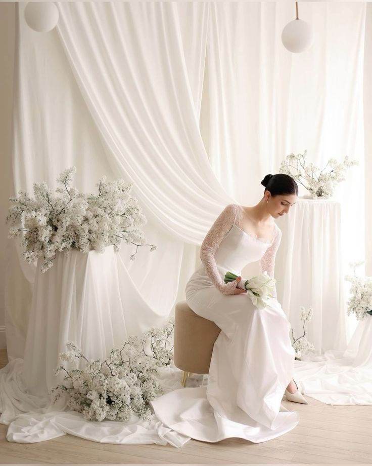 Elegant bridal scene with flowing white dress and delicate floral arrangements against draped white curtain backdrop.