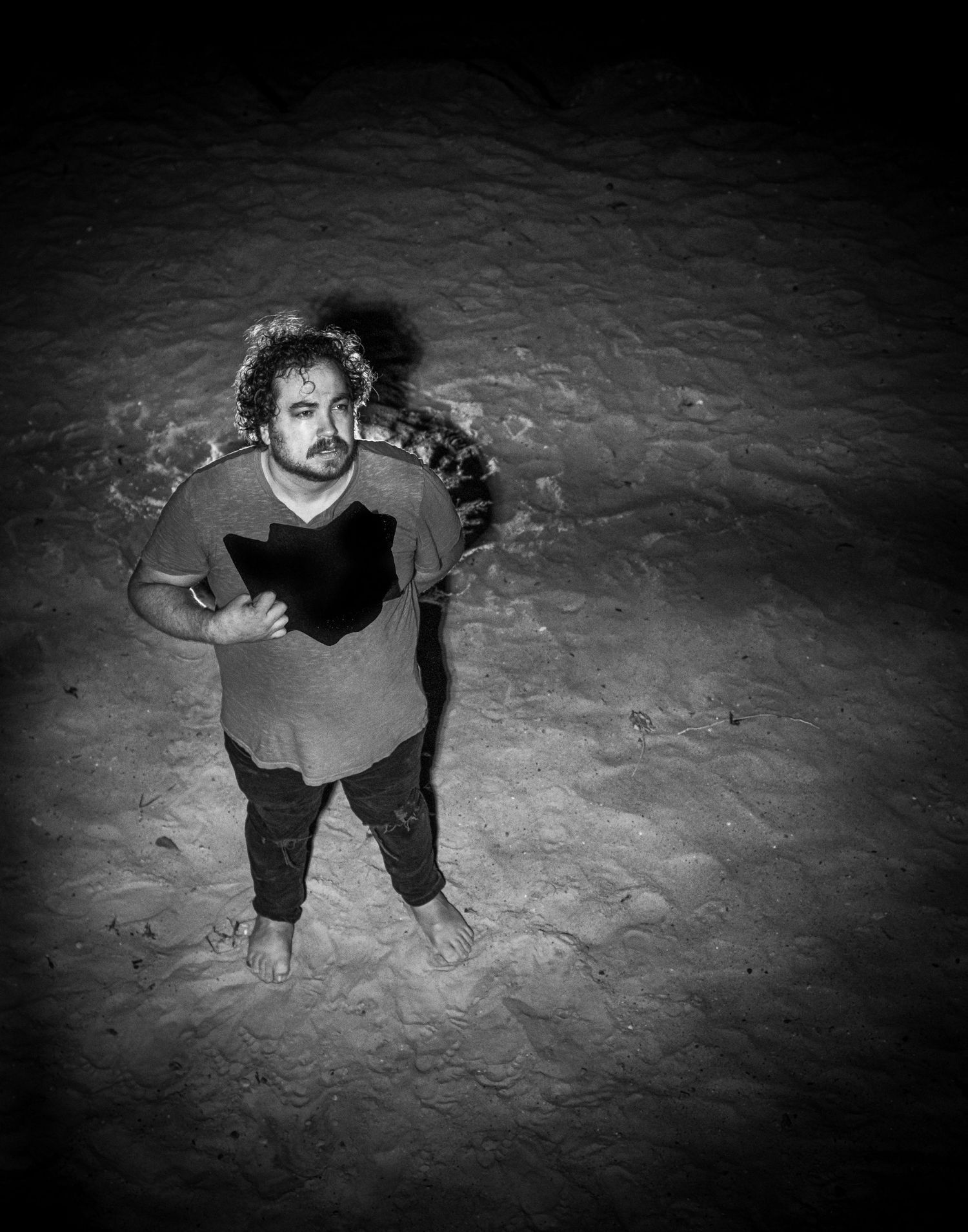 A figure stands alone on sand at night, illuminated by dramatic lighting in a black and white photograph.
