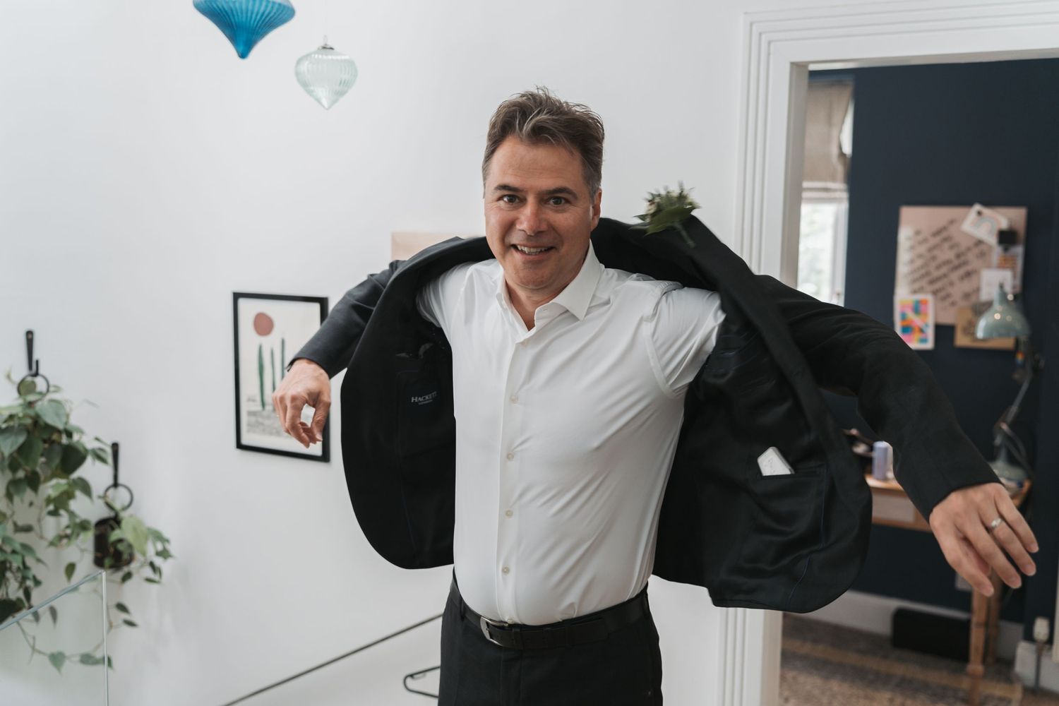 A person in a white dress shirt trying on a black suit jacket in a bright room with decor on the walls.