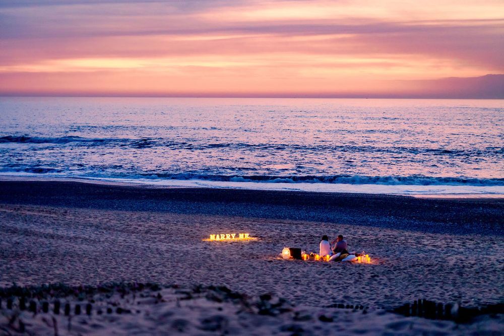 People gather around a cozy beach bonfire at sunset as pink and purple hues paint the ocean horizon.