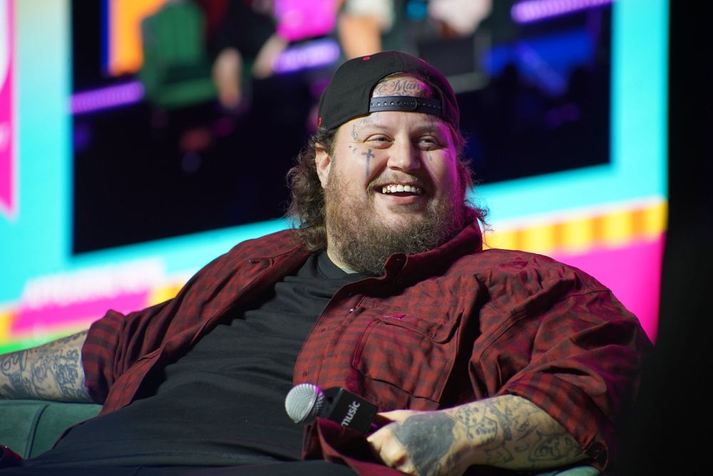 A performer in a burgundy jacket and backwards cap sits relaxed on stage during a show.