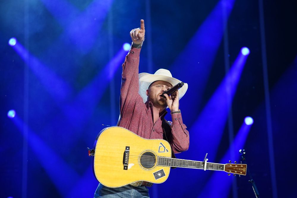 A country music performer in a cowboy hat plays acoustic guitar under blue stage lights.