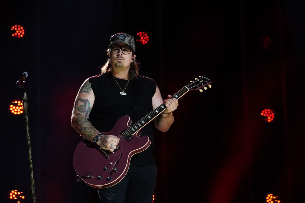 Musicians performing with guitars on dark stage with red lighting effects.