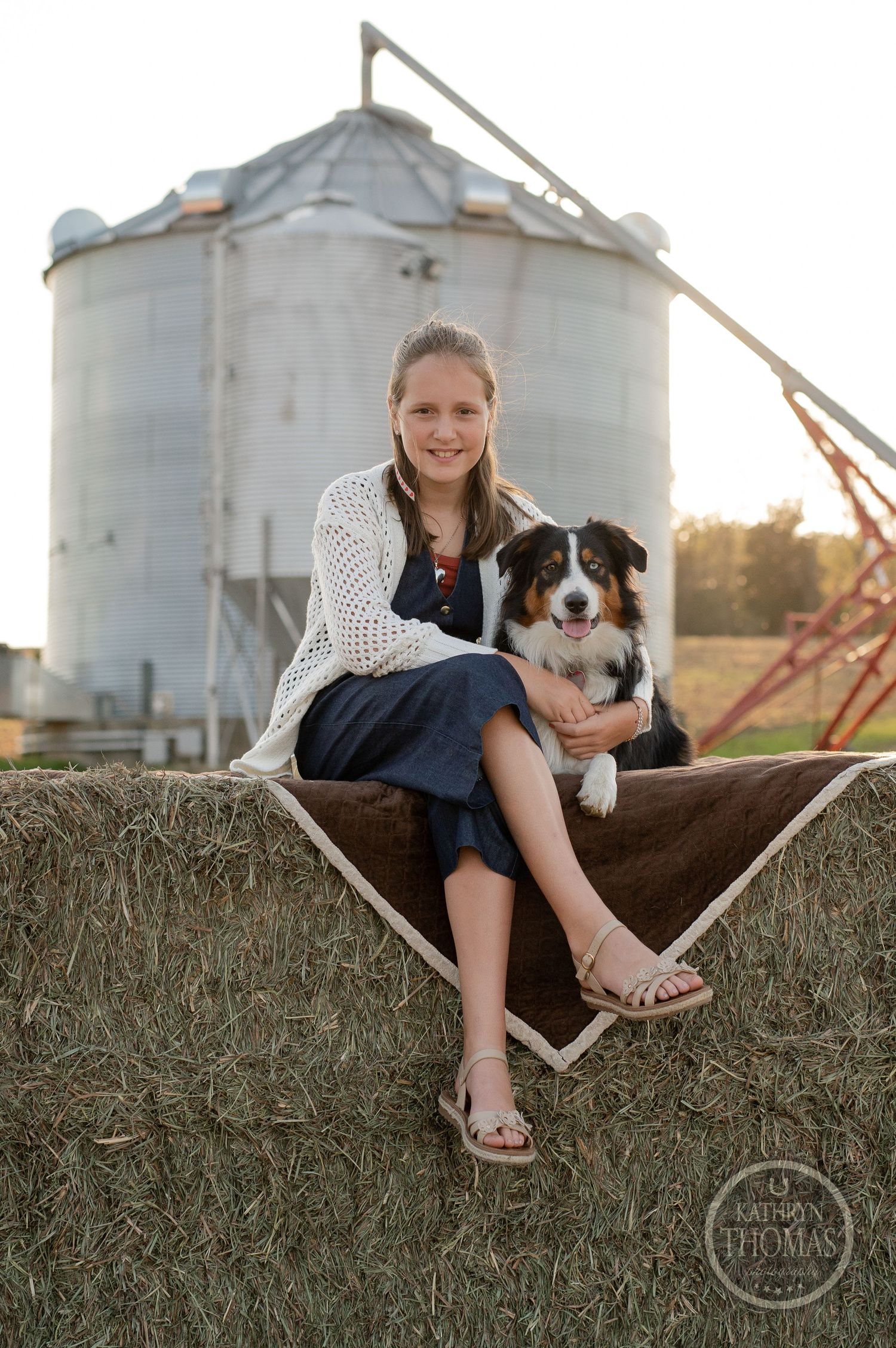 Person and Australian Shepherd sitting together on hay bale with grain silo in rural farm setting.
